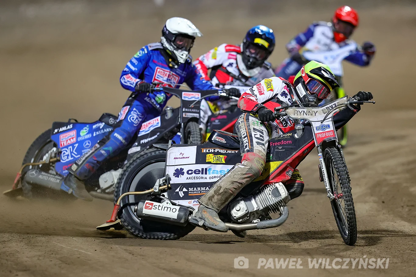 Rywalizacja czterech zawodników podczas wyścigu żużlowego na torze, motocykle nachylone w zakręcie, zawodnicy ubrani w kolorowe kombinezony i kaski, widoczne unoszące się kurz i napięcie sportowe.