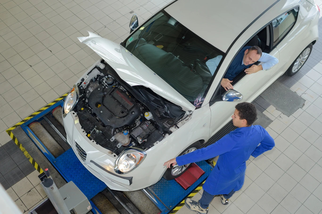 Nie wszystkie stacje kontroli pojazdów mierzą się z kryzysem. Biały samochód z otwartą maską stoi na stanowisku diagnostycznym, mechanik w niebieskim kombinezonie wykonuje badanie techniczne pojazdu, a kierowca siedzi wewnątrz auta i obserwuje przebieg testu.