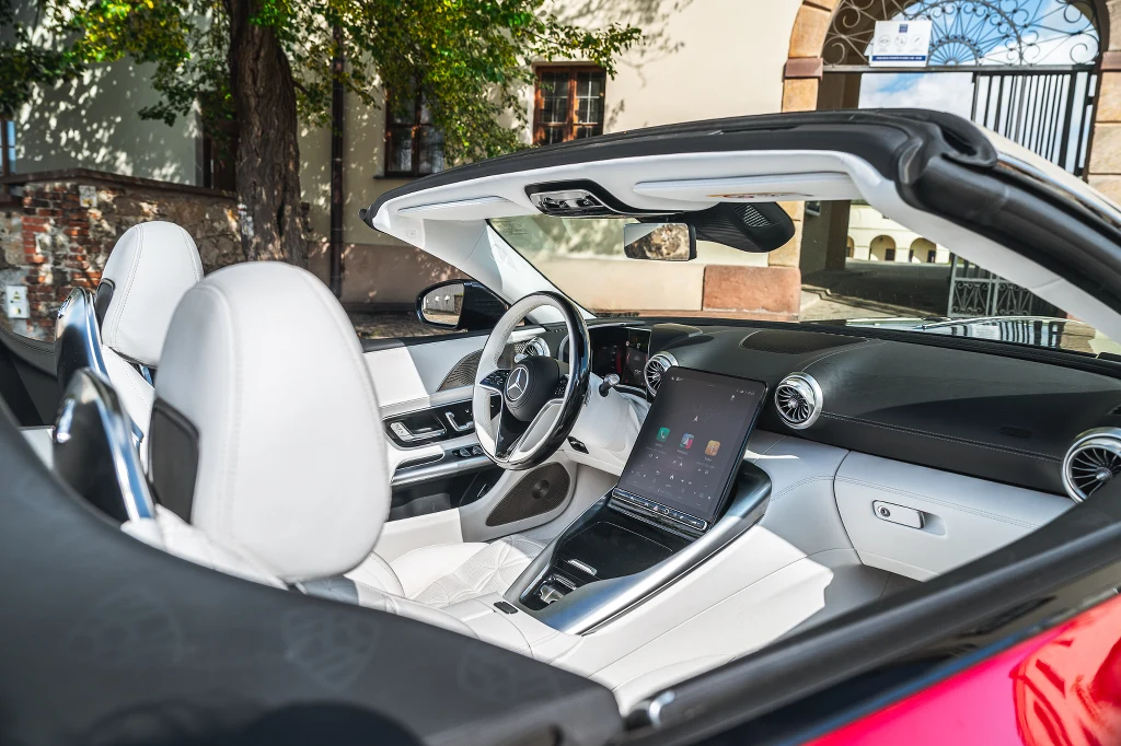 Luksusowe wnętrze samochodu kabrioletu z białą, skórzaną tapicerką, dużym panelem dotykowym na konsoli centralnej, przejrzystą deską rozdzielczą i widocznym, nowoczesnym kierownicą. Dach otwarty, a pojazd stoi na tle budynku z widocznym drzewem.