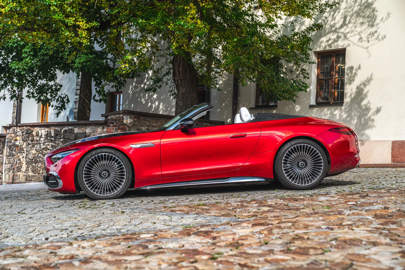 Czerwony kabriolet z opuszczonym dachem zaparkowany na kamienistej alejce przed jasnym budynkiem, otoczony zielonymi drzewami. Samochód prezentuje nowoczesny, sportowy styl i elegancję.