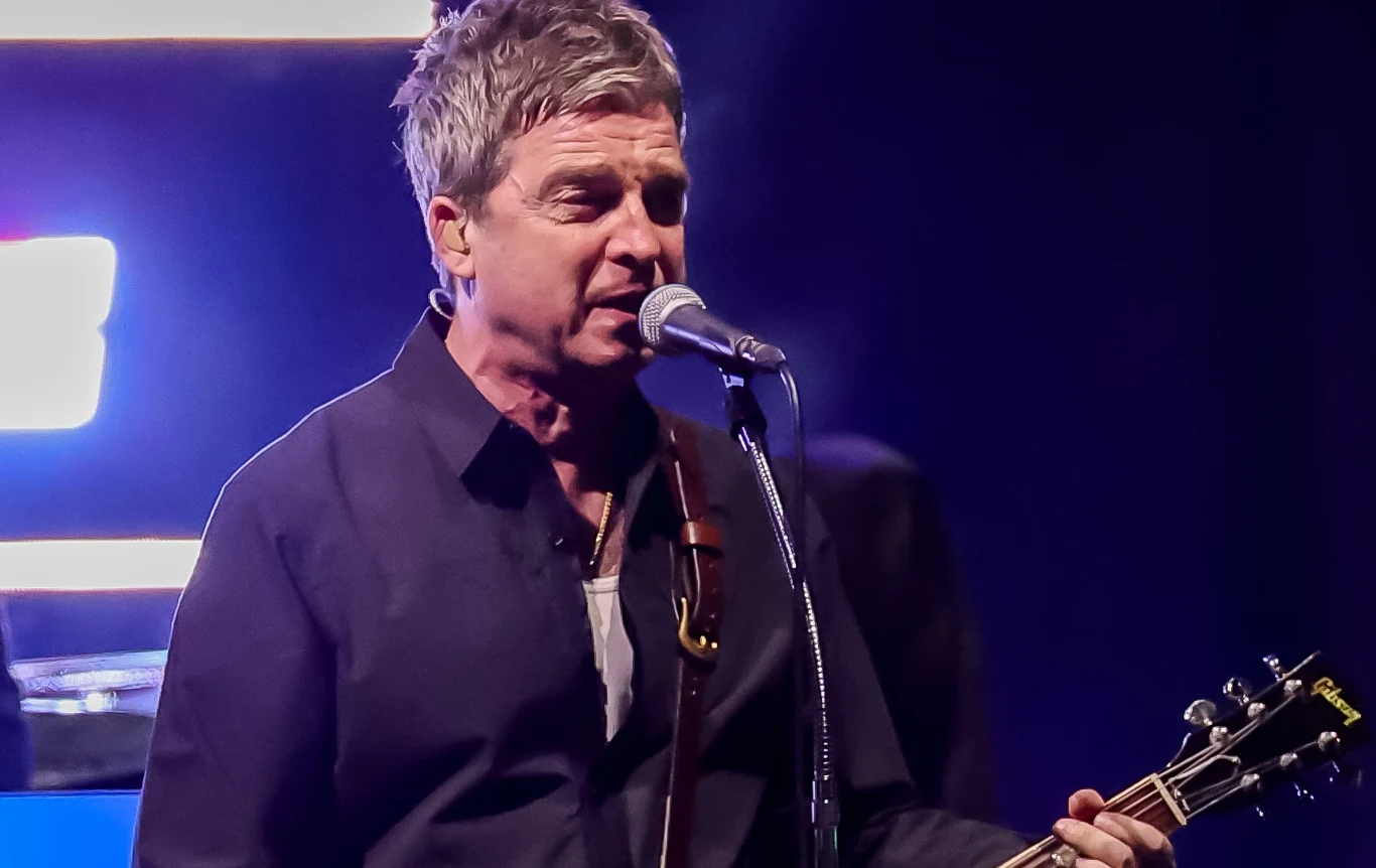 NOel Gallagher połączył soły z marką Gibson Mężczyzna śpiewający do mikrofonu podczas koncertu, trzyma gitarę elektryczną, w tle widoczne kolorowe światła sceniczne.