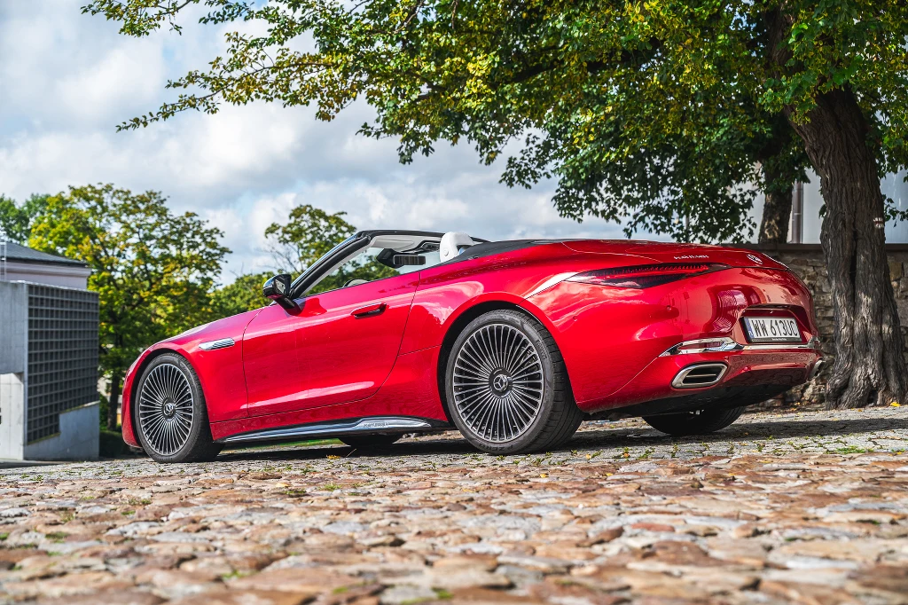 Czerwony, sportowy kabriolet Mercedes zaparkowany na brukowanej ulicy, otoczony zielenią drzew i częściowo oświetlony promieniami słońca.