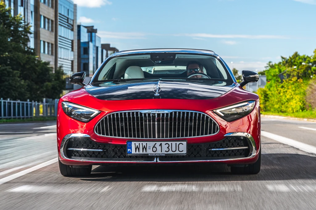Czerwony luksusowy sedan poruszający się ulicą miasta, widoczny centralnie z przodu, wyróżniający się dużą chromowaną osłoną chłodnicy, nowoczesnymi reflektorami i wypukłą maską, w tle nowoczesne budynki oraz zieleń.