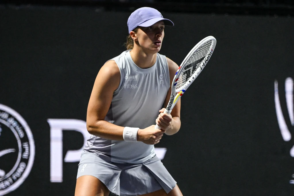 Iga Świątek podczas WTA Finals 2025 Tenisistka w sportowym stroju przygotowująca się do odbicia piłki podczas meczu, skoncentrowana, z rakietą w gotowości, ciemne tło i elementy kortu tenisowego widoczne w tle.