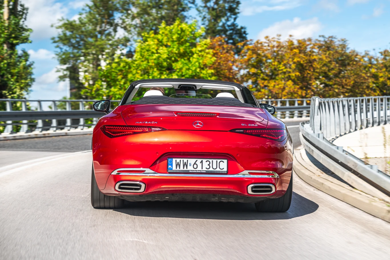 Czerwony kabriolet marki Mercedes jedzie po łuku asfaltowej drogi otoczonej barierkami, w tle drzewa z liśćmi w letnich oraz jesiennych barwach i niebo z delikatnymi chmurami.