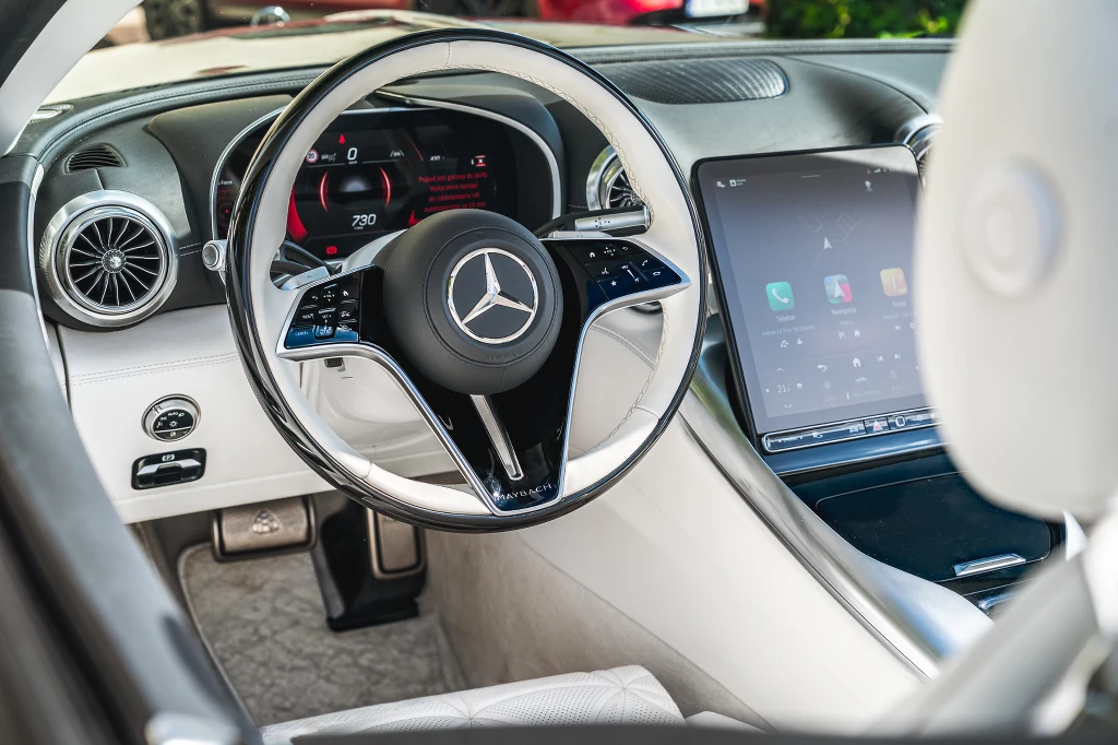 Nowoczesna kierownica z logo Mercedesa, cyfrowy wyświetlacz za kierownicą, duży ekran dotykowy po prawej stronie, jasna, elegancka tapicerka oraz elementy chromowane i wentylacyjne, akcentujące luksusowy charakter pojazdu.
