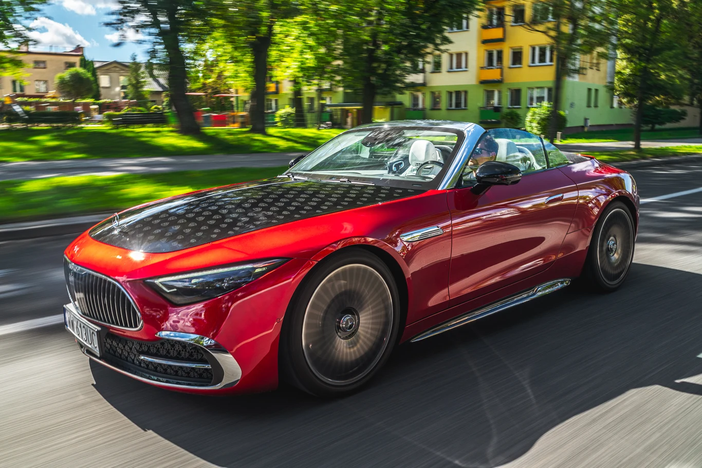 Czerwony kabriolet luksusowej marki jedzie ulicą wśród zielonych drzew i budynków mieszkalnych, dach samochodu jest złożony, wnętrze jasne, nowoczesny design i połyskująca karoseria zwracają uwagę.