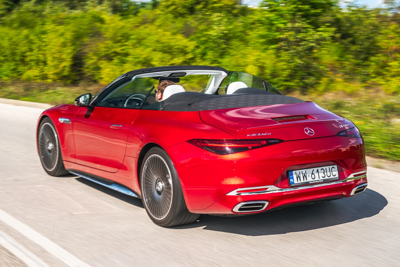 Czerwony kabriolet Mercedes-AMG SL jadący po asfaltowej drodze, otwarte dach, widoczny kierowca i pasażerowie, otoczenie zielonych krzewów w tle.