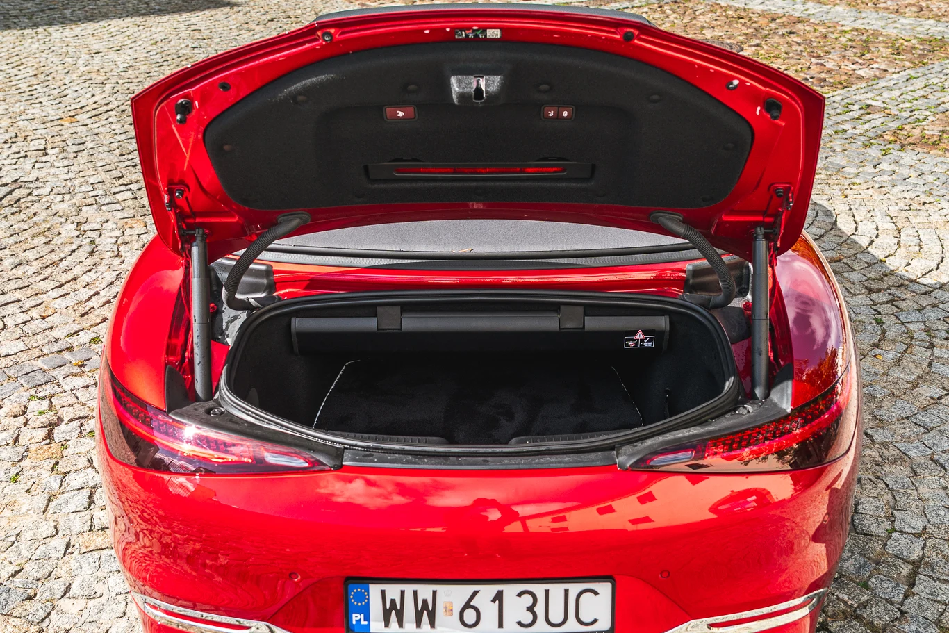 Otwarte puste bagażnik luksusowego czerwonego samochodu osobowego zaparkowanego na kostce brukowej, dobrze widoczny numer rejestracyjny oraz detale wnętrza bagażnika.