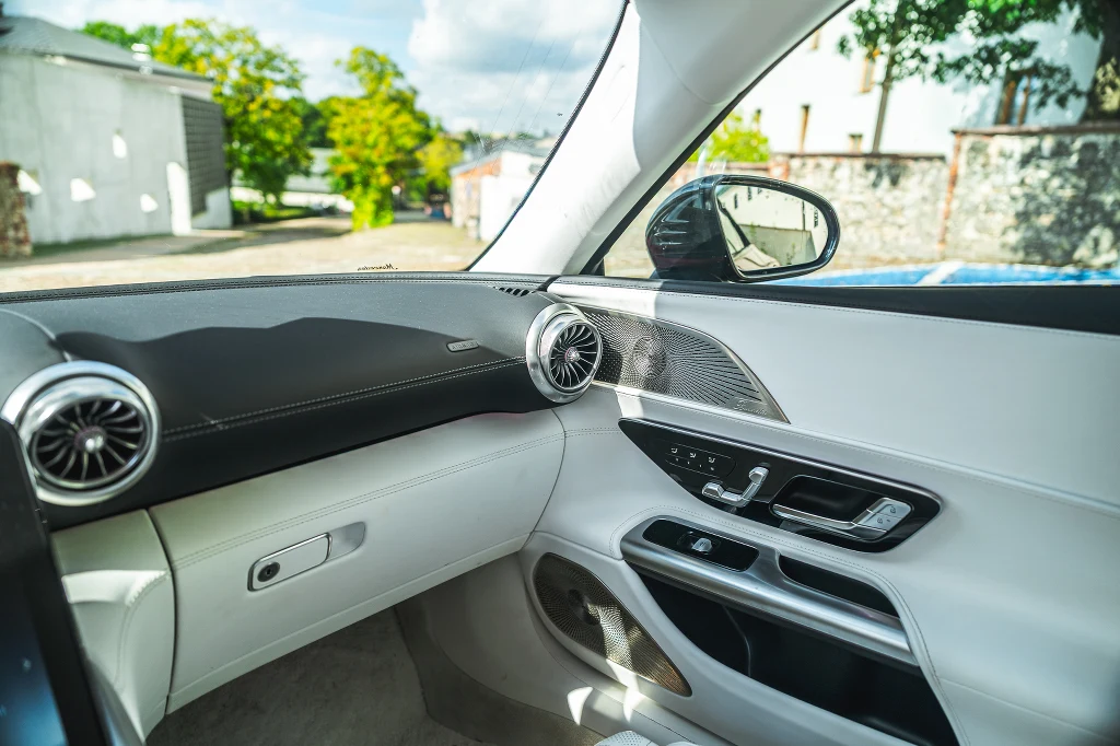 Jasne, nowoczesne wnętrze samochodu z widoczną deską rozdzielczą, okrągłymi nawiewami powietrza, eleganckimi panelami drzwiowymi i elementami sterującymi przy oknie kierowcy. Przez okno widać fragment otoczenia i zielone drzewa.
