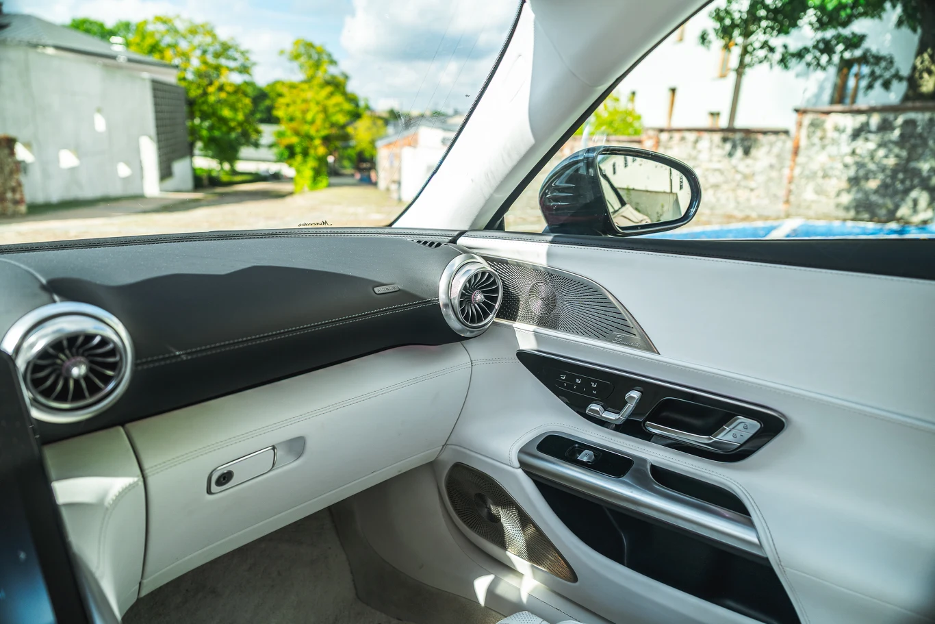 Jasne, nowoczesne wnętrze samochodu z widoczną deską rozdzielczą, okrągłymi nawiewami powietrza, eleganckimi panelami drzwiowymi i elementami sterującymi przy oknie kierowcy. Przez okno widać fragment otoczenia i zielone drzewa.