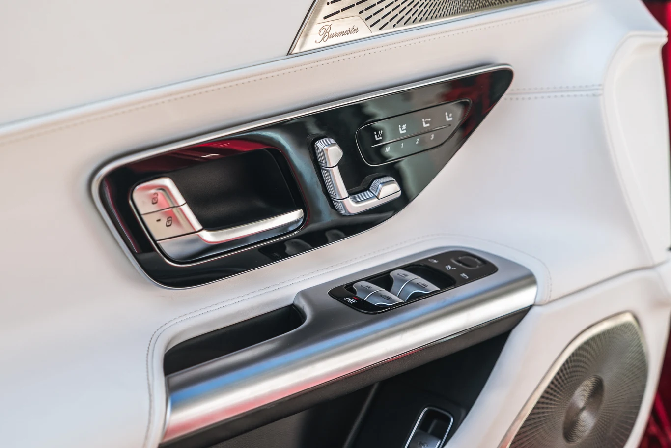 Panel drzwi samochodu wykończony jasną skórą, z widocznym zestawem przycisków do regulacji szyb, foteli i lusterka oraz głośnikiem audio.