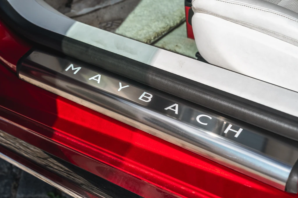 Próg wejściowy luksusowego samochodu z widocznym napisem 'Maybach' na metalowej listwie oraz fragment białej skórzanej tapicerki i karoserii w kolorze czerwonym.