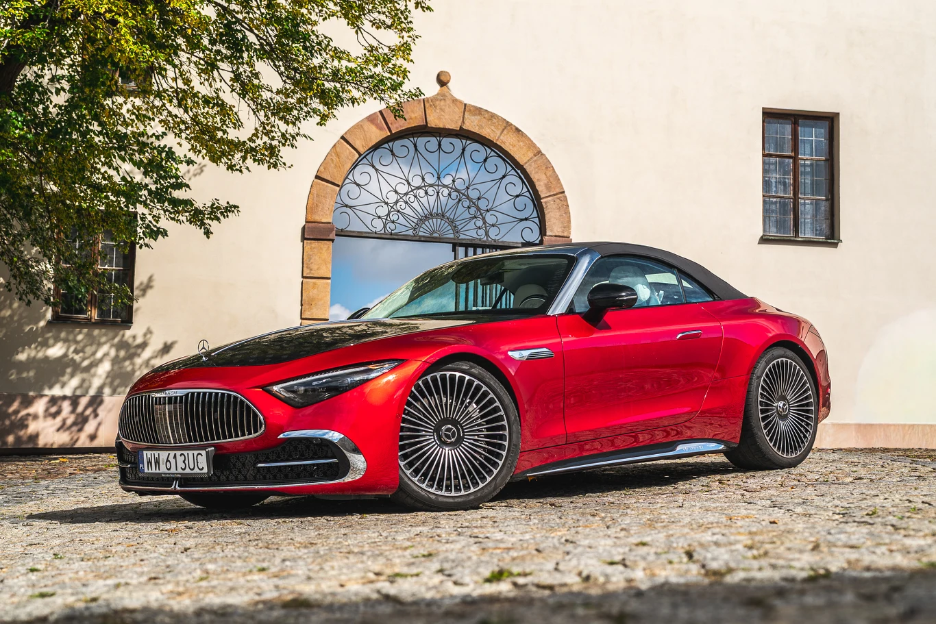 Czerwony sportowy kabriolet zaparkowany na brukowanym dziedzińcu przed elegancką kamienicą z ozdobnym łukiem nad drzwiami i oknem, auto wyróżnia się dynamicznym designem i stylowymi felgami.