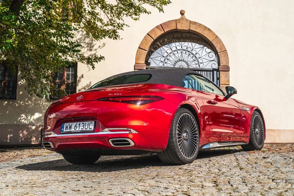 Czerwony kabriolet Mercedes-Benz AMG SL stoi na brukowanym podjeździe przed stylową bramą z kutego żelaza, w tle jasna ściana budynku oraz cień rzucany przez drzewo.