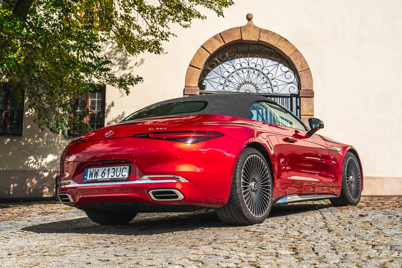 Czerwony kabriolet Mercedes-Benz AMG SL stoi na brukowanym podjeździe przed stylową bramą z kutego żelaza, w tle jasna ściana budynku oraz cień rzucany przez drzewo.