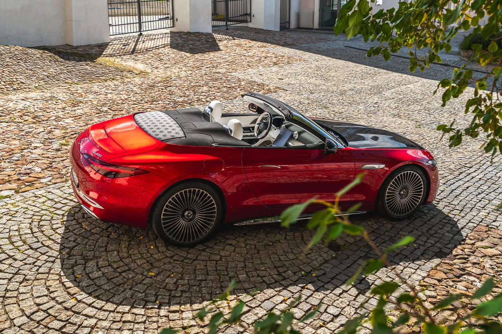 Czerwony kabriolet klasy premium z opuszczanym dachem stoi zaparkowany na brukowanym dziedzińcu przy jasnym budynku, wokół widoczne są liście drzew i charakterystyczne wzory na kołach pojazdu.