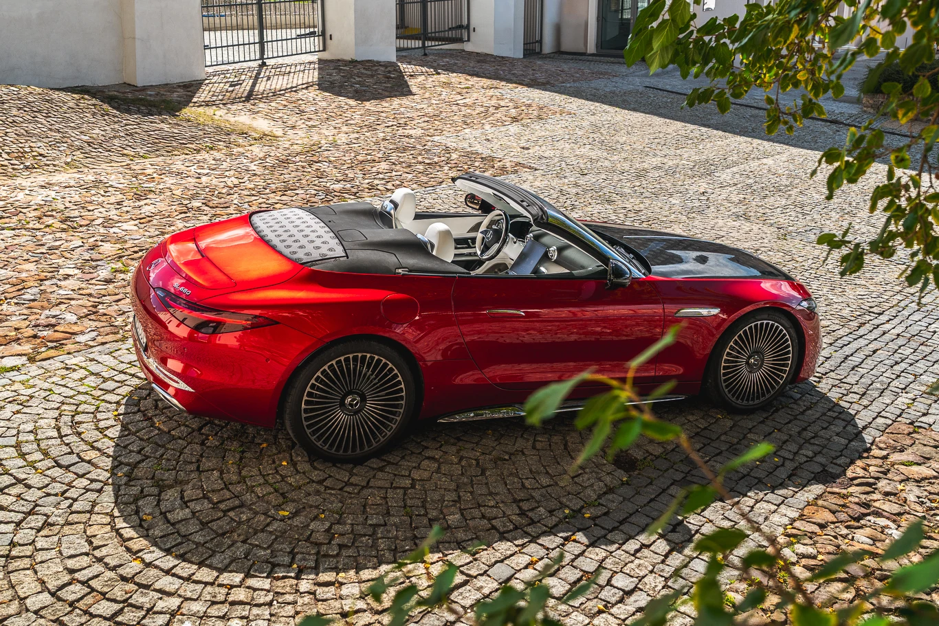 Czerwony kabriolet klasy premium z opuszczanym dachem stoi zaparkowany na brukowanym dziedzińcu przy jasnym budynku, wokół widoczne są liście drzew i charakterystyczne wzory na kołach pojazdu.