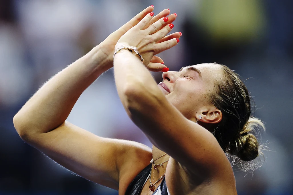 Sportowiec o jasnych włosach z zamkniętymi oczami i splecionymi dłońmi pod brodą, wyraża silne emocje, prawdopodobnie ulgę lub wzruszenie po zakończeniu meczu. W tle rozmyte sylwetki publiczności.