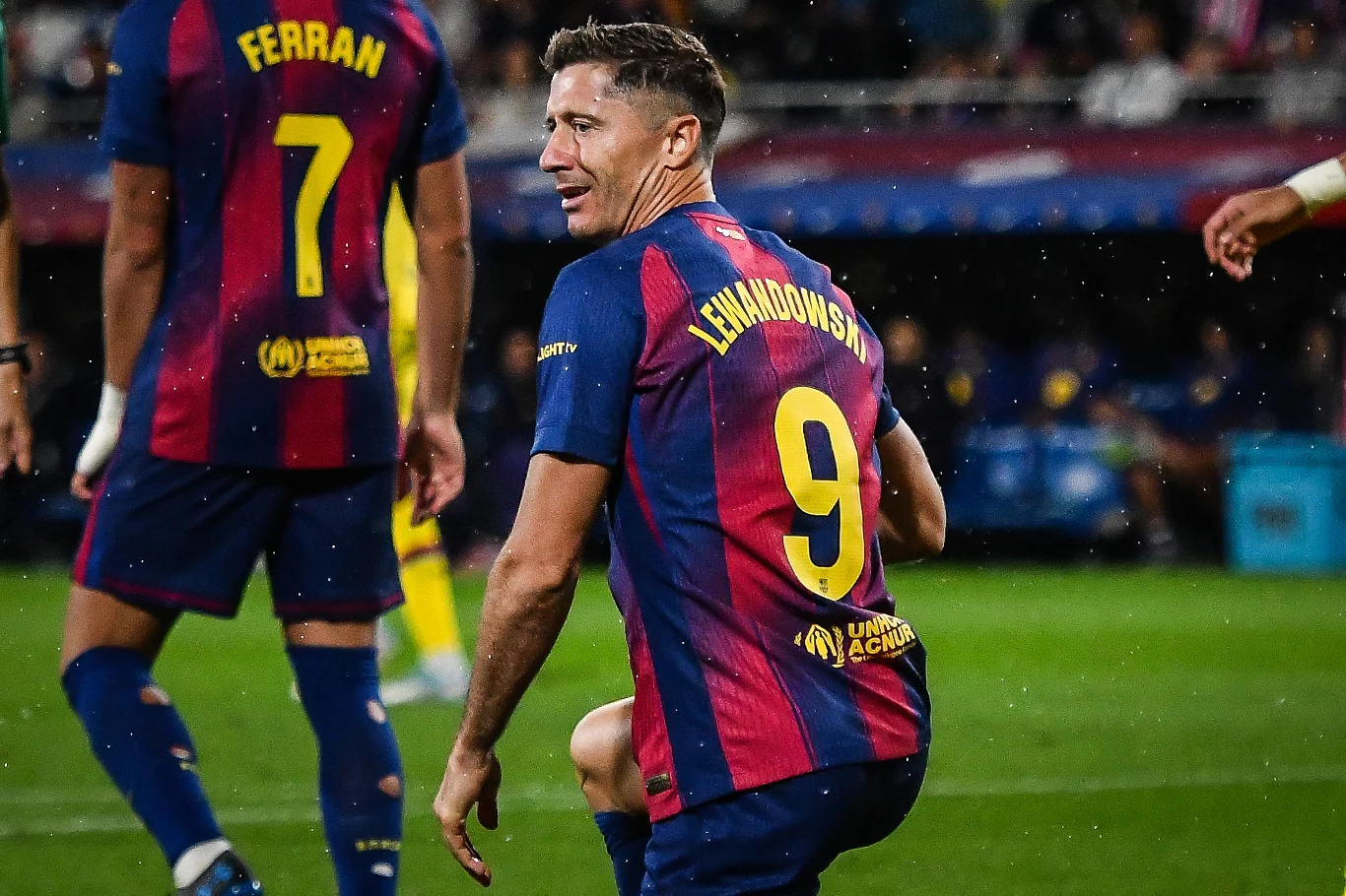 Robert Lewandowski Piłkarz w stroju FC Barcelony z numerem 9 i nazwiskiem Lewandowski na plecach klęczy na murawie, obok widoczny drugi zawodnik z numerem 7 Ferran, w tle rozmyty stadion i kibice.