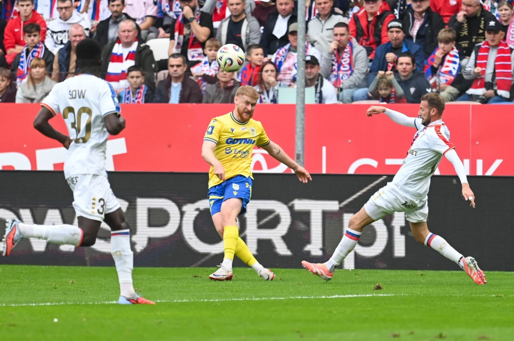 Kadr meczu Górnik Zabrze - Arka Gdynia Dwóch piłkarzy walczy o piłkę podczas meczu piłkarskiego, widownia na trybunach uważnie obserwuje przebieg spotkania, jeden zawodnik kopie piłkę w kierunku bramki.