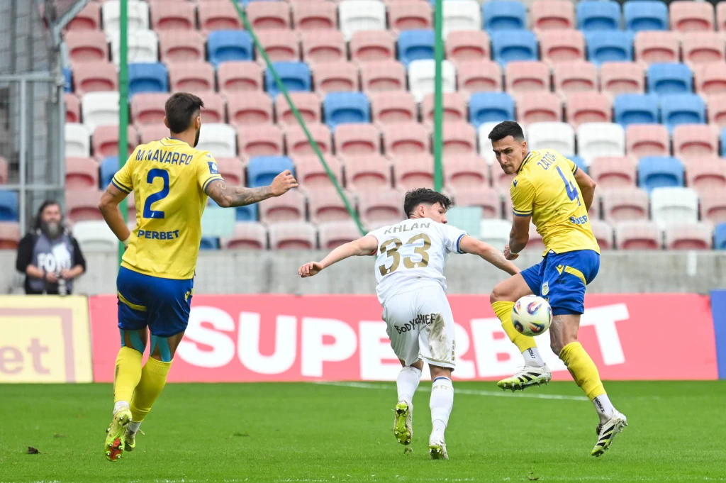 Kadr meczu Górnik Zabrze - Arka Gdynia Piłkarze w trakcie intensywnej akcji na boisku, jeden zawodnik w białym stroju próbuje odebrać piłkę dwóm zawodnikom w żółto-niebieskich strojach na tle trybun stadionu.