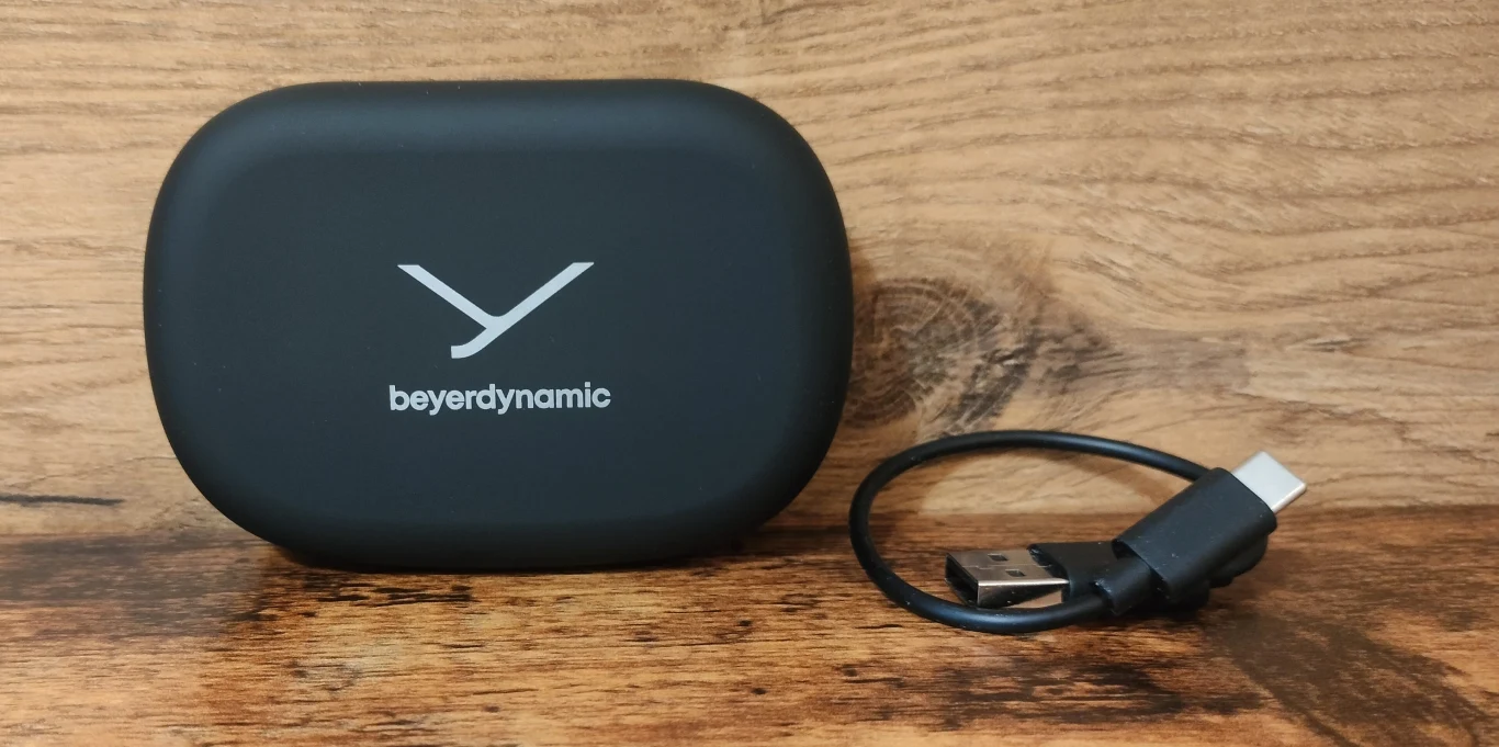 Beyerdynamic Verio 200 Czarna, zaokrąglona etui z nadrukowanym logotypem beyerdynamic ustawiona na drewnianej powierzchni, obok krótki kabel USB typu C.