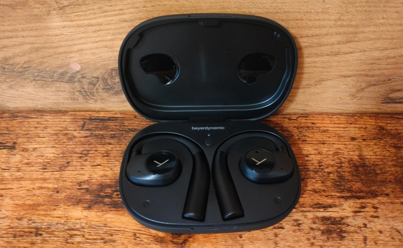 Beyerdynamic Verio 200 Czarne bezprzewodowe słuchawki douszne w etui ładującym, umieszczone na drewnianej powierzchni. Widoczne logo producenta na słuchawkach i wnętrzu etui.