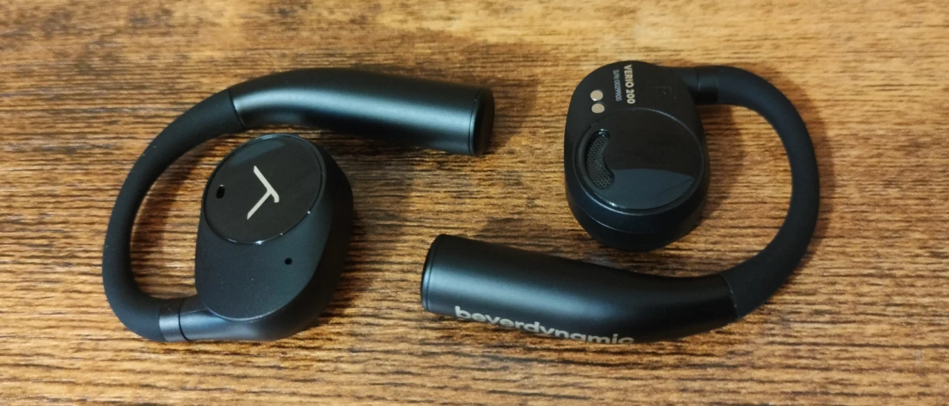 Beyerdynamic Verio 200 Czarne bezprzewodowe słuchawki douszne z pałąkiem na ucho leżą na drewnianej powierzchni. Widoczne logo producenta oraz przyciski na jednej ze słuchawek.