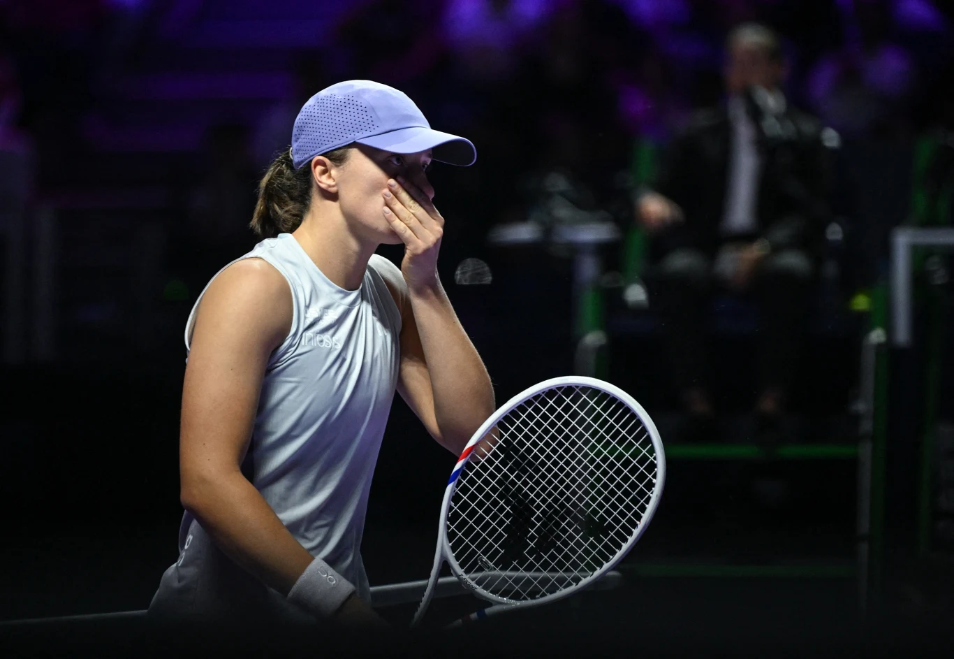 Iga Świątek Tenisistka ubrana w sportowy strój i czapkę z daszkiem stoi na korcie trzymając rakietę, zakrywając usta dłonią, w tle rozmyta sylwetka sędziego i subtelne oświetlenie.