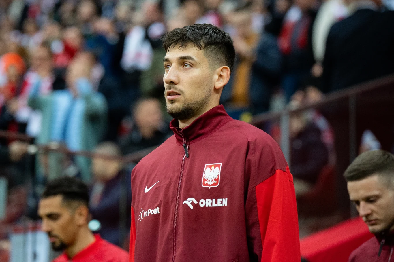 Piłkarz w czerwonym dresie reprezentacji Polski, stojący wśród innych zawodników na stadionie. W tle rozmyty tłum kibiców na trybunach.