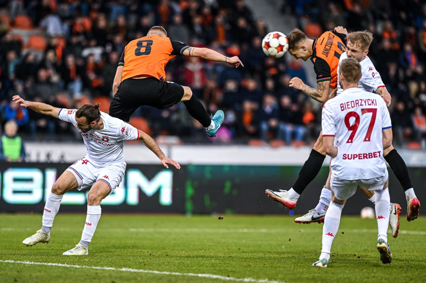 W meczu Chrobry Głogów - Wisła Kraków nie brakowało walki Piłkarze dwóch drużyn walczą o piłkę w powietrzu podczas meczu piłki nożnej, jeden z zawodników skacze wysoko starając się główkować, a pozostali próbują przejąć kontrolę nad futbolówką, w tle widoczna publiczność na trybunach.