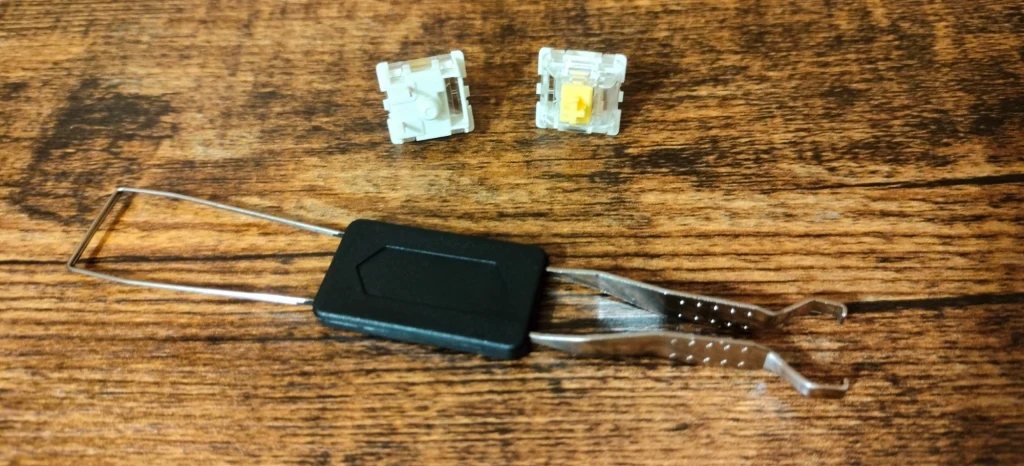 Genesis Thor 404 Metalowy klucz do wyjmowania przełączników mechanicznych do klawiatur oraz dwa osobne przełączniki z przezroczystej obudowy, leżące na drewnianej powierzchni.