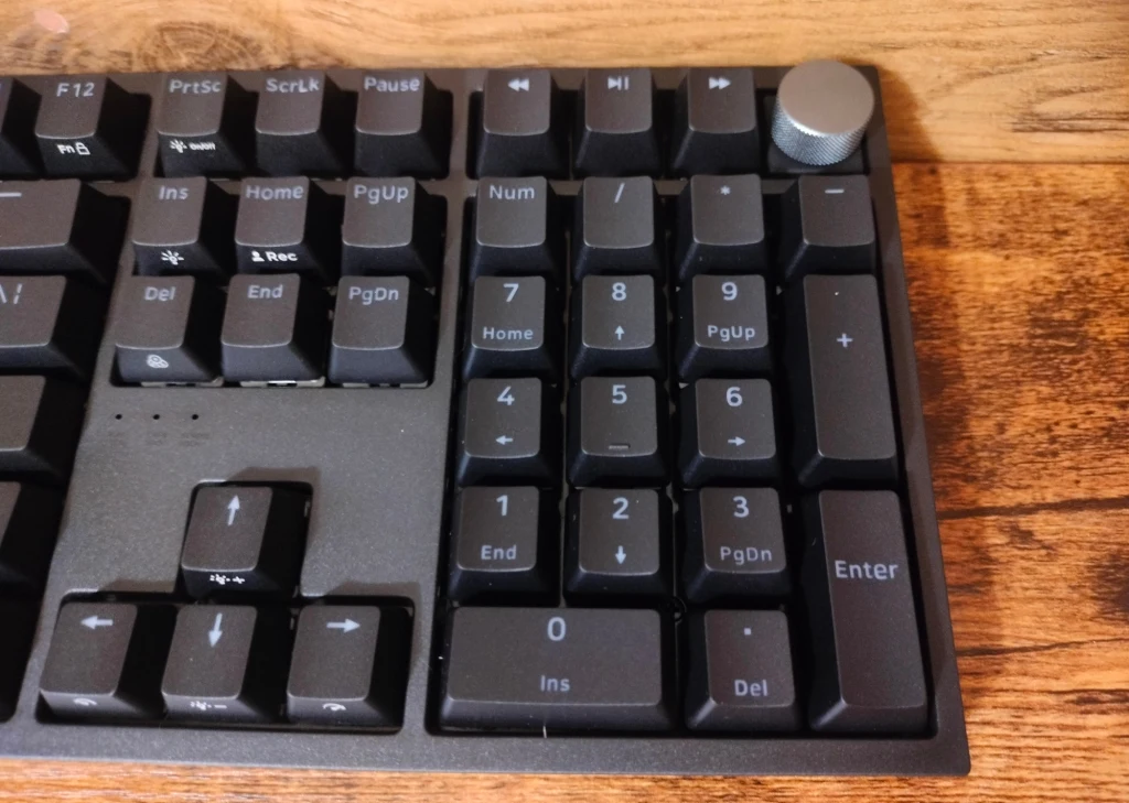 Genesis Thor 404 Fragment klawiatury komputerowej z sekcją numeryczną, klawiszami funkcyjnymi i pokrętłem ulokowanym w prawym górnym rogu, leżącej na drewnianym biurku.