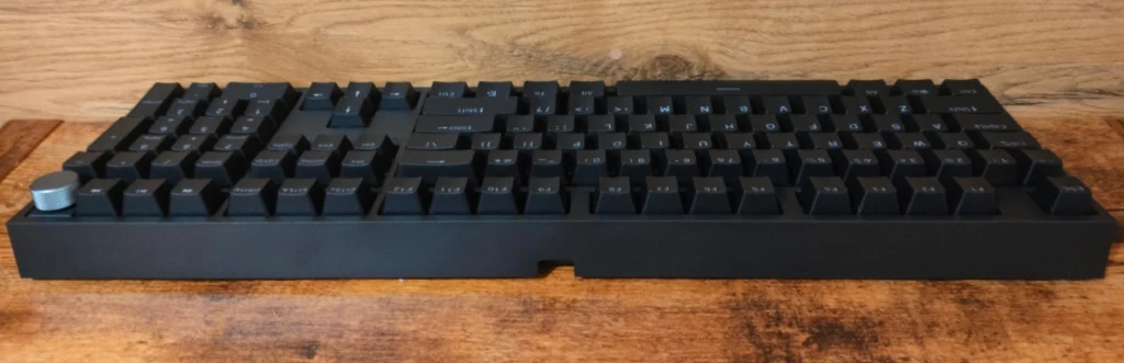 Genesis Thor 404 Czarna klawiatura mechaniczna ustawiona na drewnianym blacie z tłem w postaci drewnianej ściany, wyposażona w dodatkowe pokrętło w lewym górnym rogu.