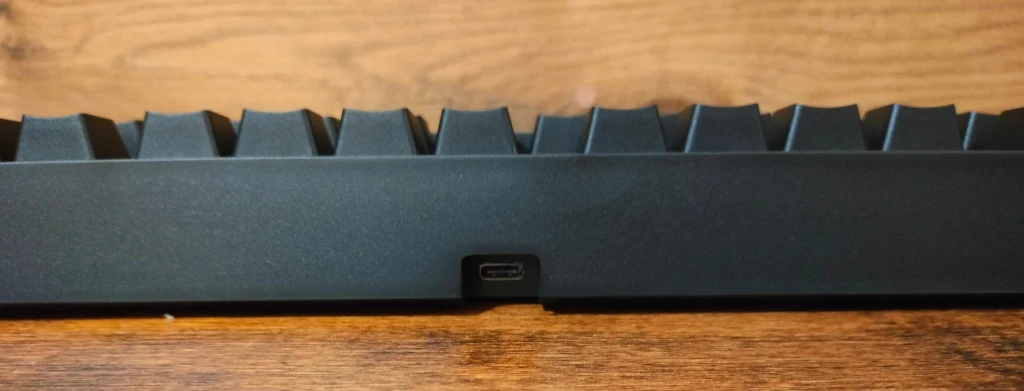 Genesis Thor 404 Fragment czarnej klawiatury mechanicznej na tle drewnianego biurka, z widocznym portem USB-C pośrodku dolnej krawędzi obudowy.