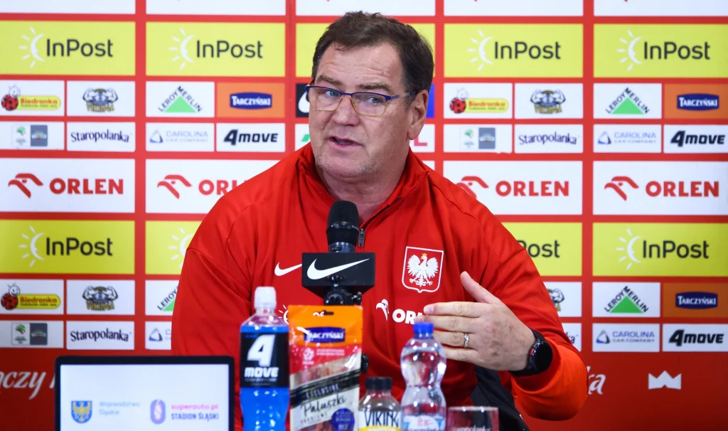 Mężczyzna w czerwonej bluzie z orzełkiem na piersi siedzi przy stole konferencyjnym, mówi do mikrofonu, w tle tablica z logotypami sponsorów.