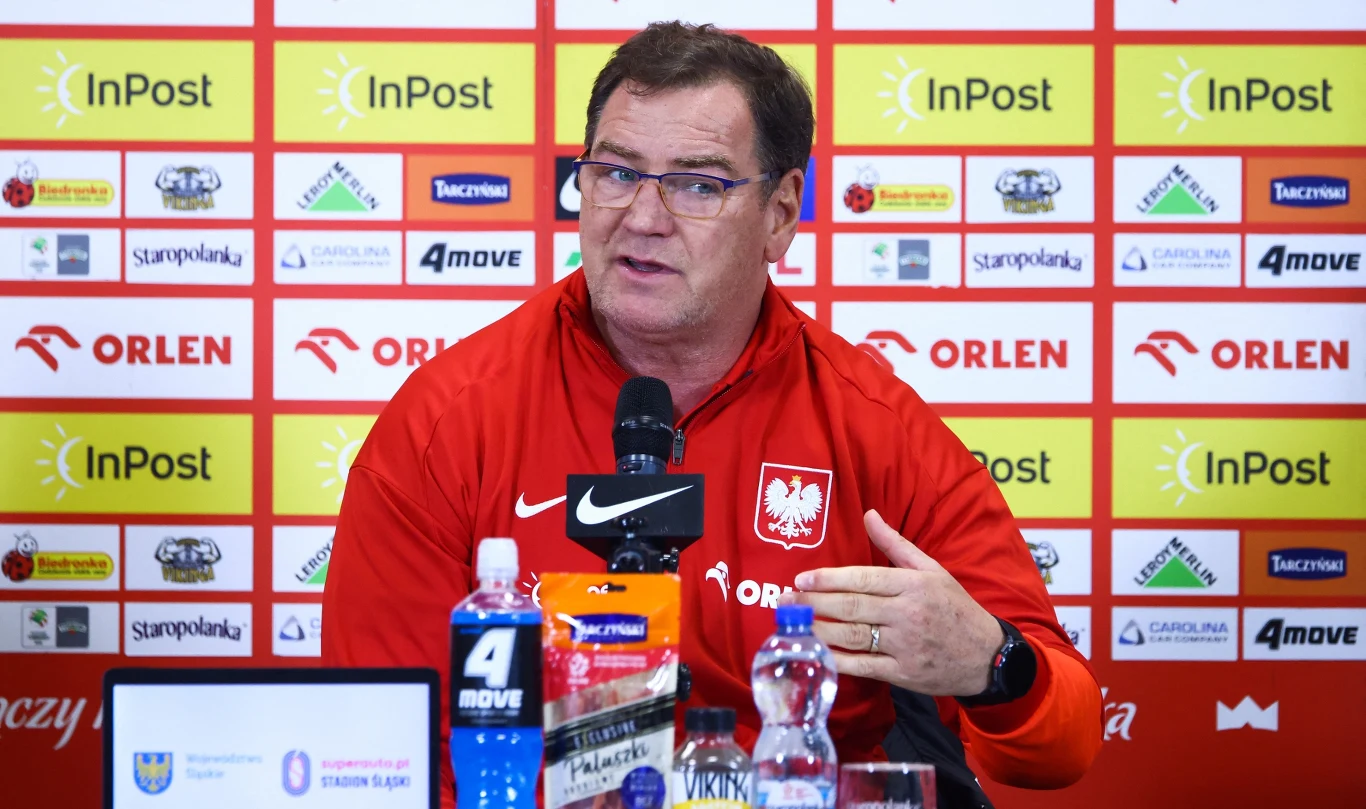 Mężczyzna w czerwonej bluzie z orzełkiem na piersi siedzi przy stole konferencyjnym, mówi do mikrofonu, w tle tablica z logotypami sponsorów.