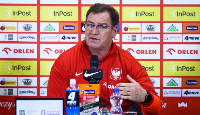 Mężczyzna w czerwonej bluzie z orzełkiem na piersi siedzi przy stole konferencyjnym, mówi do mikrofonu, w tle tablica z logotypami sponsorów.