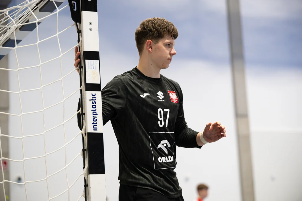 Dominik Tylman, bramkarz reprezentacji Polski U-18 Młody mężczyzna w czarnym stroju bramkarza piłki ręcznej z numerem 97 i godłem Polski na piersi stoi przy bramce na hali sportowej, trzymając się słupka.