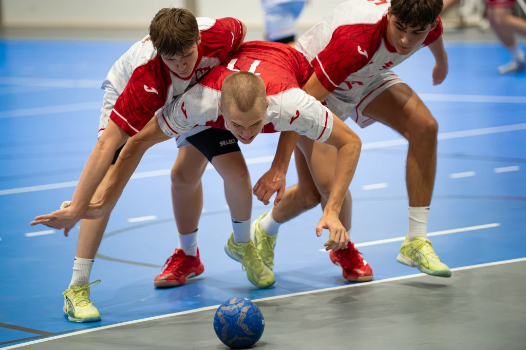 Mecz reprezentacji Polski prowadzonej przez Rafała Glińskiego Trzech młodych mężczyzn w sportowych strojach walczy o piłkę na hali, skupiając się na dynamicznej rywalizacji podczas meczu piłki ręcznej.