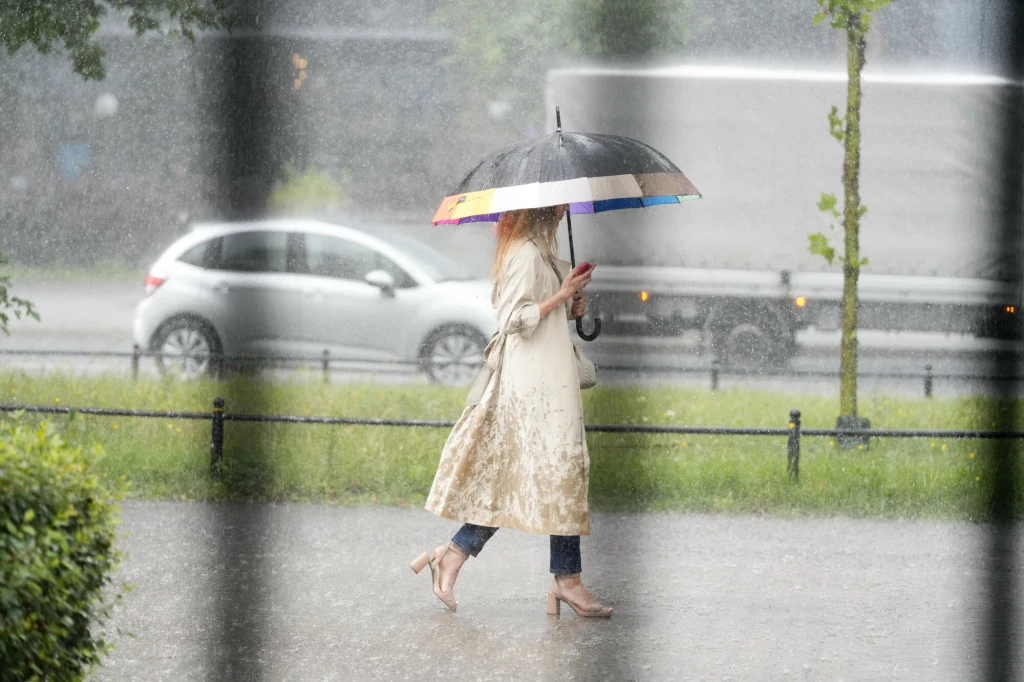 Nadciąga załamanie pogody (zdj. ilustracyjne) Kobieta w jasnym płaszczu i sandałach przechodzi przez ulicę podczas intensywnego deszczu, trzymając w ręku kolorowy parasol i telefon. W tle widoczne są zaparkowane samochody i zieleń miejska.