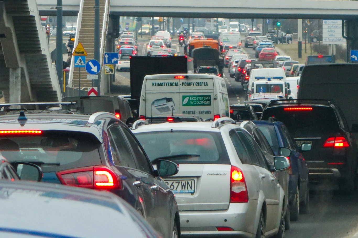 Samochody stojące w gęstym korku na zatłoczonej ulicy miasta, widocznych wiele różnych pojazdów, most i elementy miejskiej infrastruktury drogowej.