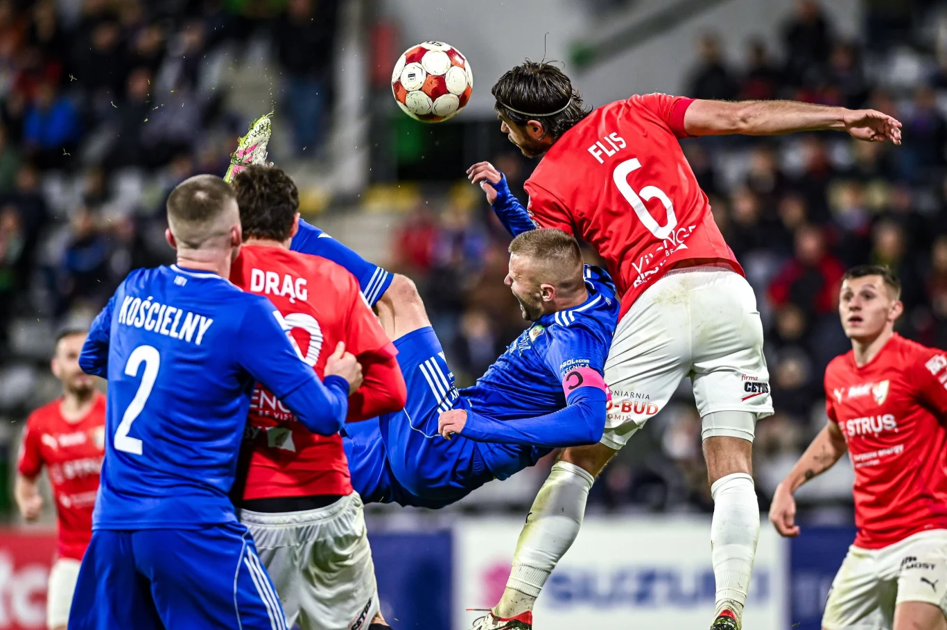 Piłkarze dwóch drużyn rywalizują o piłkę podczas meczu, jeden z zawodników próbuje przewrotki, a reszta graczy walczy w powietrzu w polu karnym. W tle rozmazana publiczność i trybuny stadionu.