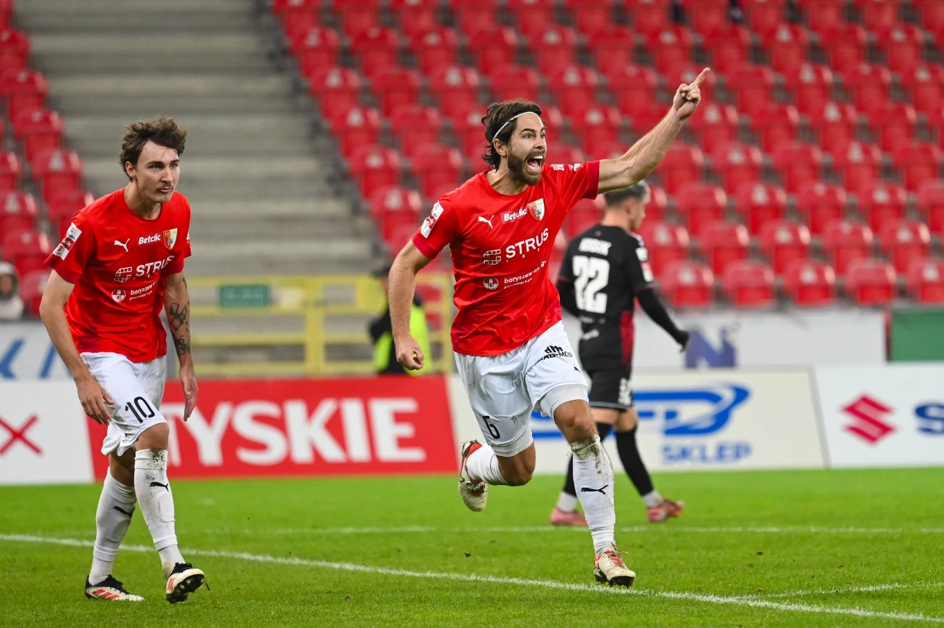 Dwóch piłkarzy w czerwonych koszulkach i białych spodenkach świętuje zdobycie gola na tle pustych trybun, zawodnik po prawej podnosi rękę w geście triumfu, z tyłu widoczni inni gracze oraz banery reklamowe.