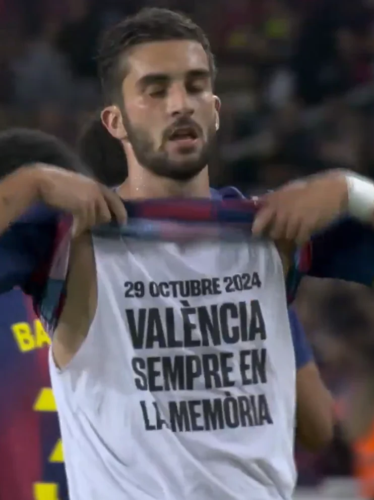 Piłkarz unosi koszulkę, pod którą znajduje się biała koszulka z napisem upamiętniającym wydarzenie z 29 października 2024 roku oraz odniesienie do miasta Walencja. W tle widać rozmyte postacie innych zawodników oraz stadion.