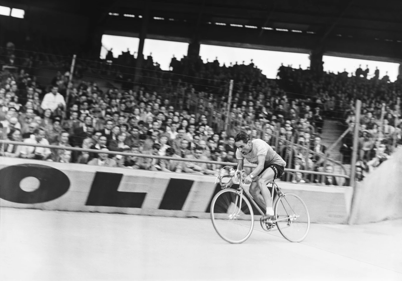 Zawodowy kolarz jedzie na torze rowerowym podczas wyścigu, wokół trybuny wypełnione widzami śledzącymi rywalizację.
