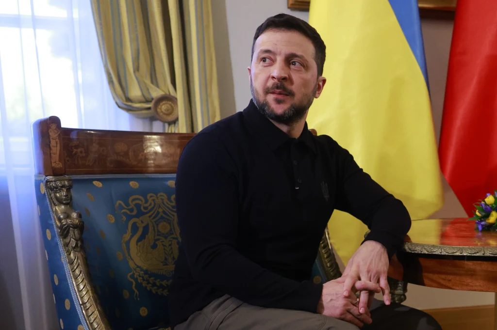 Wołodymyr Zełenski Mężczyzna w ciemnej koszuli siedzi na ozdobnym fotelu, w tle widoczne flagi Ukrainy i Polski oraz elegancki wystrój wnętrza.