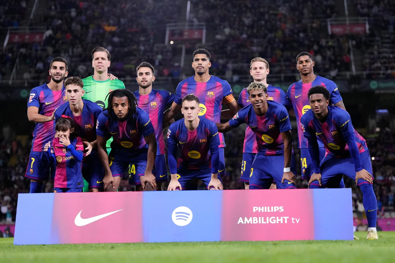 Grupa jedenastu piłkarzy w jednolitych strojach klubowych pozujących na stadionie piłkarskim przed meczem, na pierwszym planie baner z logotypami sponsorów i dużą widownią w tle.