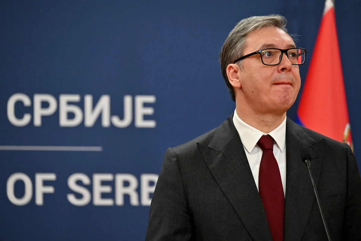 Prezydent Serbii Aleksandar Vuczić Mężczyzna w garniturze i czerwonym krawacie stoi na tle niebieskiego baneru z napisem w języku serbskim oraz flagą Serbii po prawej stronie; patrzy lekko w górę, obok znajduje się mikrofon